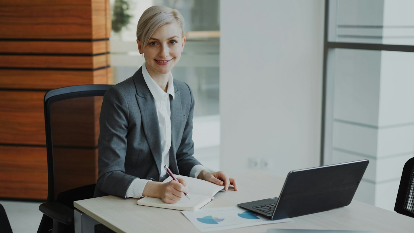 Businesswoman smiles while writing in a notebook at her desk.